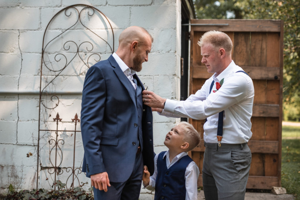 The groom's brother assists him in wearing his boutonniere as his son looks up in admiration.