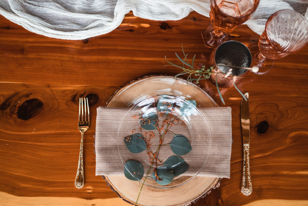 Clear plate setting with eucalyptus branch and gold silverware, copper mugs, and vintage colored glassware