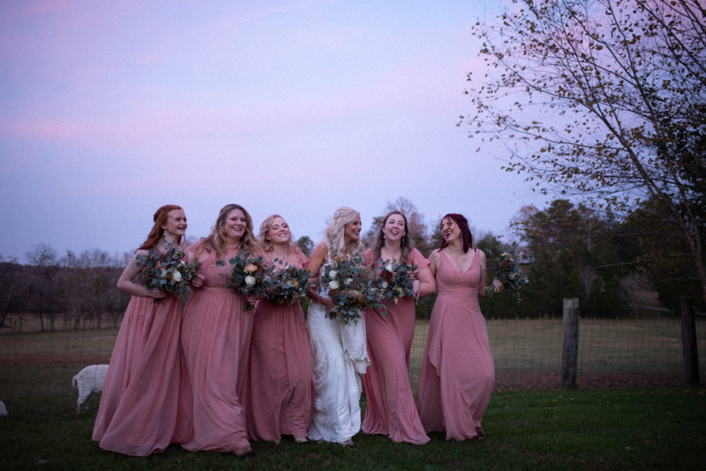 Bride and bridesmaids laugh walking arm and arm under a cotton candy sky.
