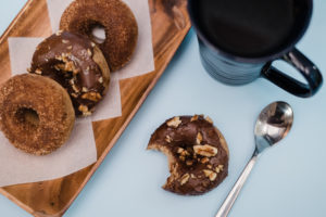 Vegan and Gluten free chocolate frosted turtle donut on blue surface with coffee in blue mug near wooden serving tray with more gluten free donuts.