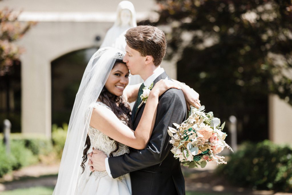 Groom kisses his new bride on the fore-head in the courtyard at st benedicts catholic church in alpharetta, ga