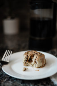 Gluten free vegan sweet cinnamon roll on a white plate with a bite taken out of it in front of coffee in a french press.