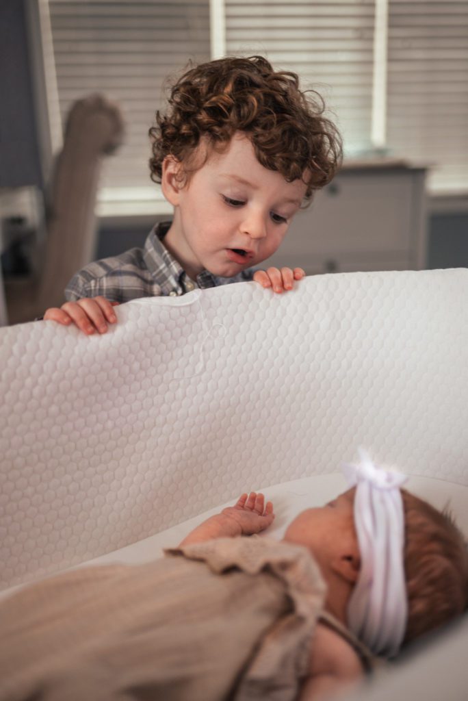 Toddler boy with brown curly hair looks over edge of bassinet to peek on his newborn sister dressed in beige ruffles and a white bow.