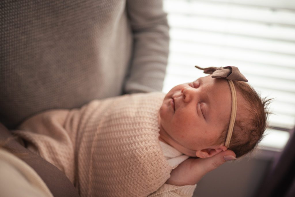 newborn baby held by a window during in home newborn photography session in st louis. Baby has large bow and is wrapped in a soft beige swaddle.
