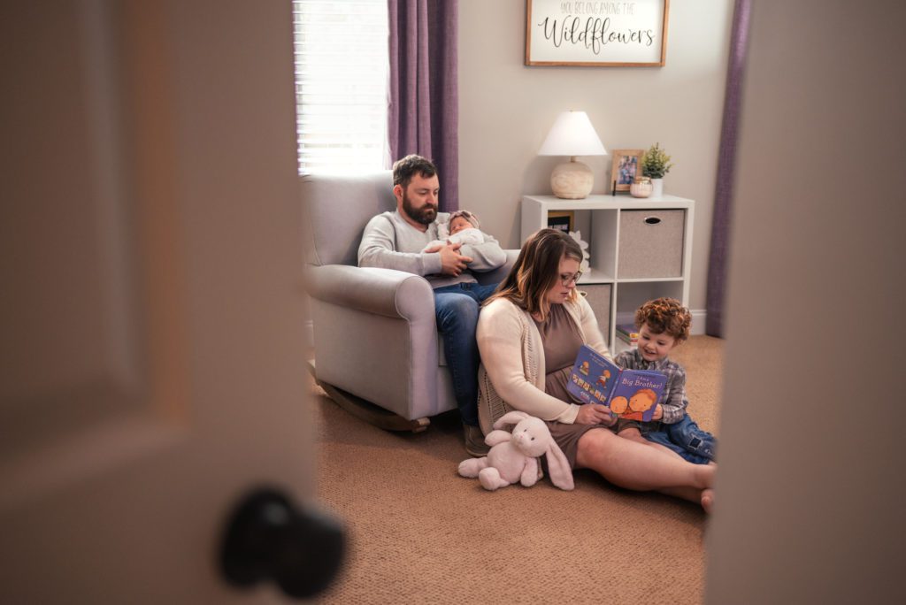 Dad hold baby girl in his arms as mom reads toddler boy all about being a big brother as they spend time together in the purple and grey nursery while the photographer peeks through the crack in the door.
