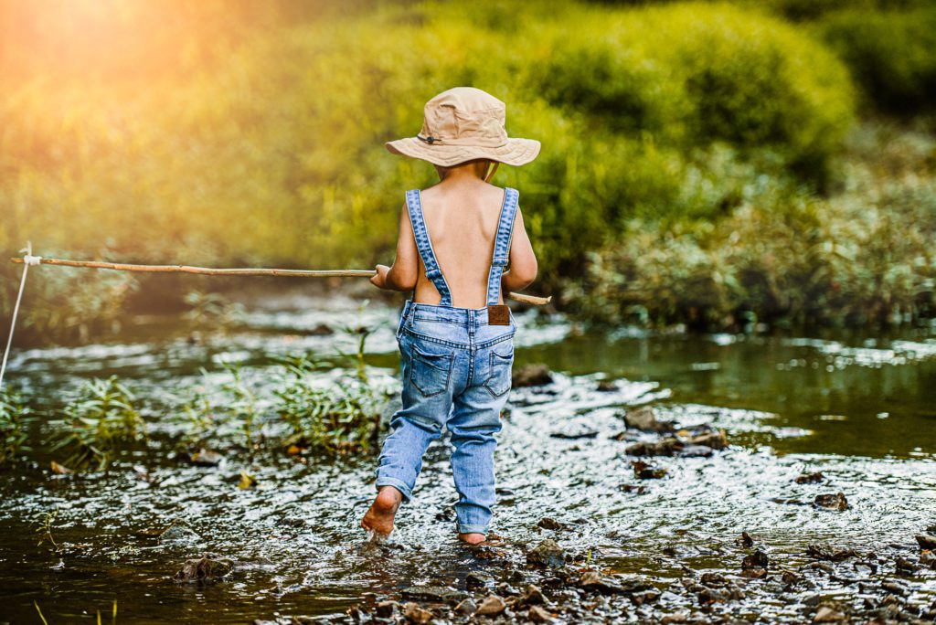 Little boy in overalls and fishing hat walk through creek at Quail Ridge park in Wentzville, Missouri with his fishing pole and fishing hat.