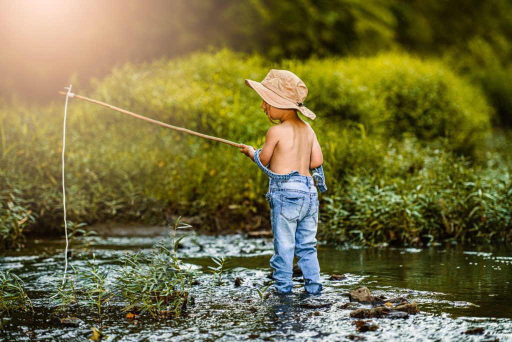 Toddle boy takes his fishing pole to the creek at golden hour. Overall straps fall off his shoulders as he stand in the creek waiting for a bit - fine art photography portrait for Children
