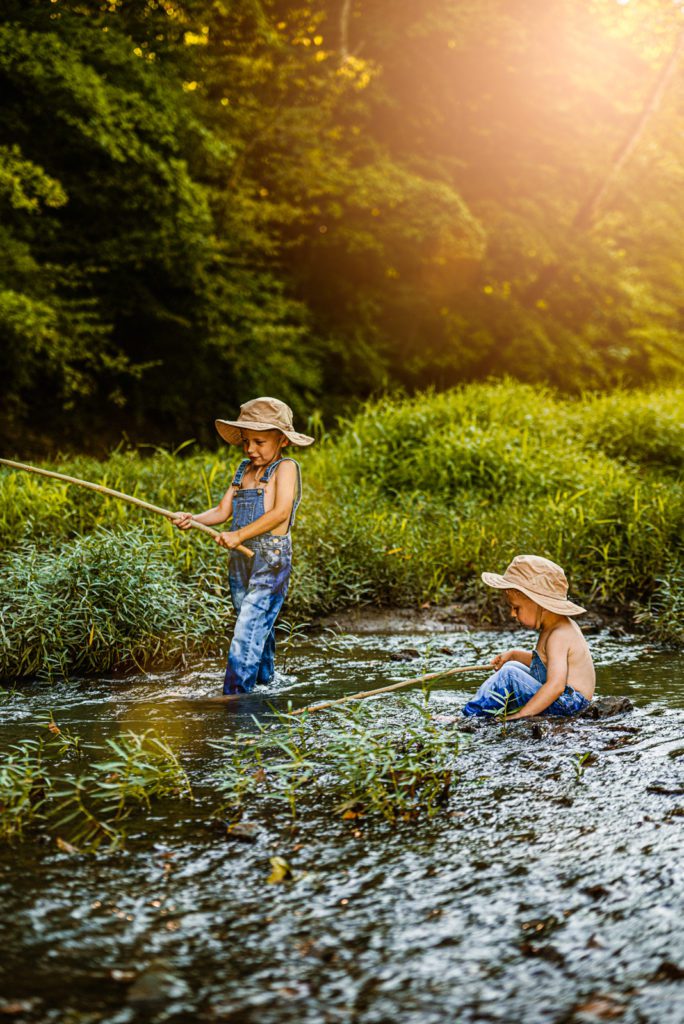 Older brother holds on tight to his fishing pole in wentzville, MO while toddler brother looks at rocks in the Creek near St Louis MO with family photographer Becky Hassel
