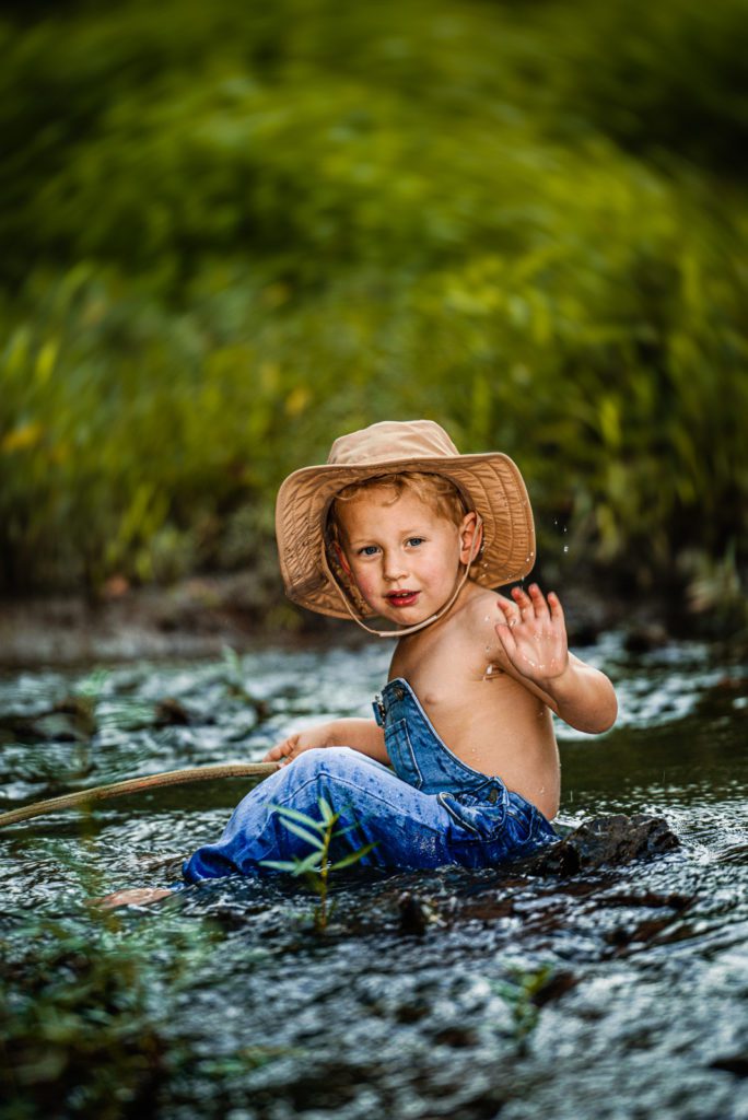 Toddler boy blonde hair blue eyes sits in creek with overalls and a fishing hat splashing in the water for a artistic family photography image in St Louis Missouri