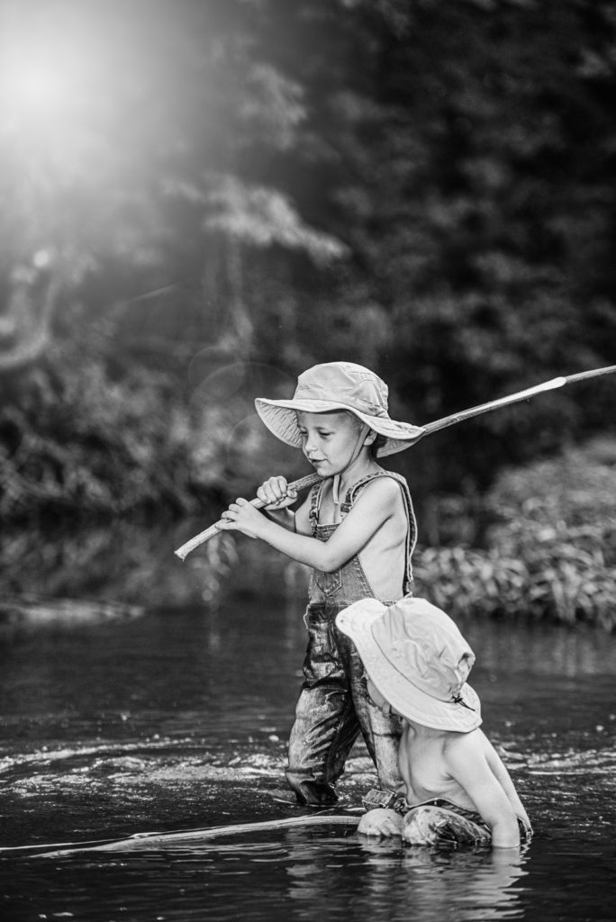 Black and white photography portrait by St Louis family photographer Becky hassel. Two young brothers wade through the creek with the fishing hats as the sun sets on them.