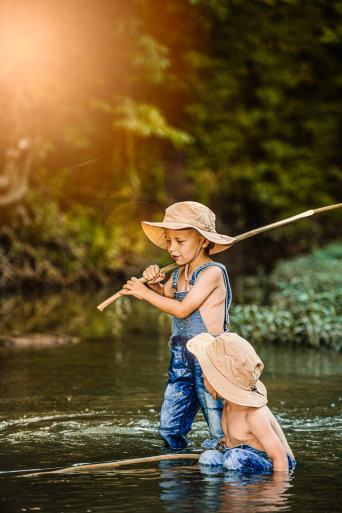 Like a minature Huck Finn and Tom Sawyer, two brothers go exploring in the creek at golden hour with their fishing poles and the sun shining through the trees creating fine art photography.