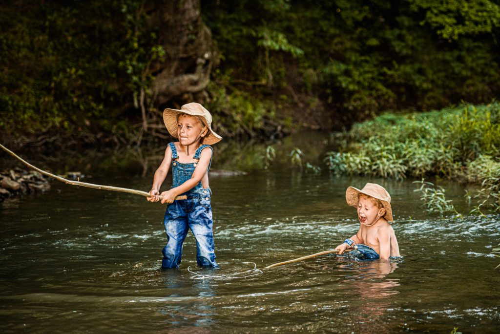 Older brother casts his pole and surprises little brother. They both laugh with joy sitting in the creek in Missouri
