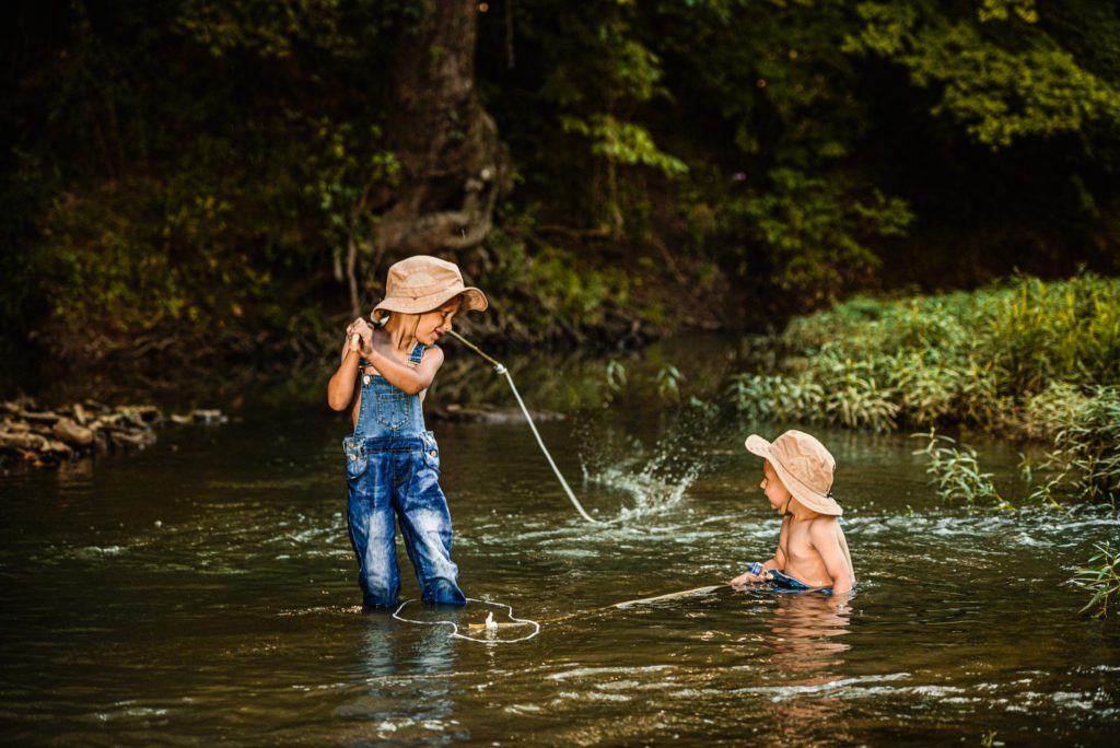 Young boy watches his fishing line as splashes behind him and his little brother watches him as he plays in the missouri creek for fun family portraits