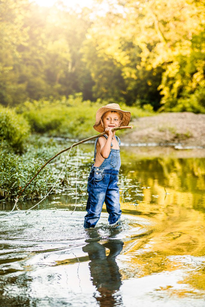 Young boy goes out flying fishing at golden hour in the creek with blue overalls and a tan fishing hat and the fishing pole over his shoulder.