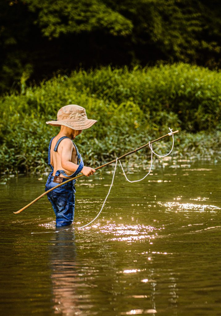 Toddler in jean overalls and fishing hat holds his fishing pole as he searches for the fish that got away in this fine art photography session at golden hour in St Louis, MO