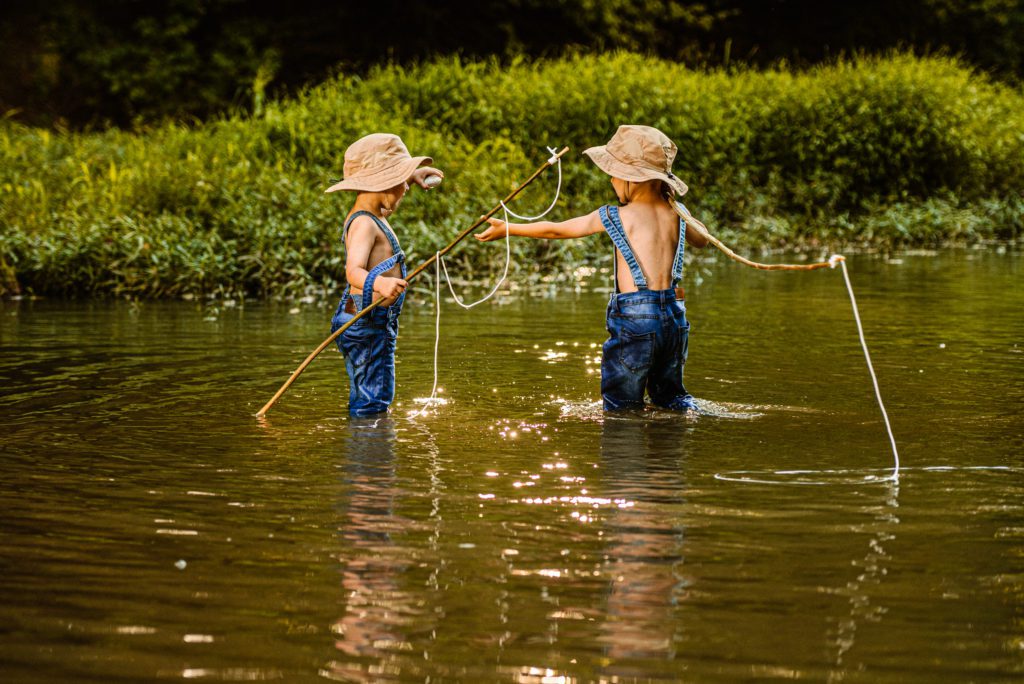 Two young boys in jean overalls and fishing hats stand in the creek as the older brother helps younger brother with his fishing pole for photos with two brothers children photography