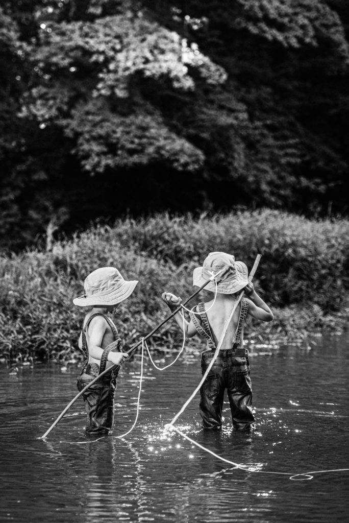 Black and white of Two young boys in overalls and and fishing hats with fishing poles prepare to throw a rock in this unique boys fishing photography session for fine art portraits
