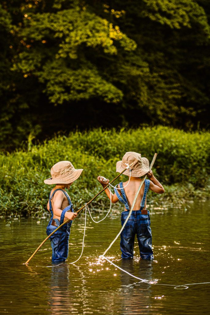 Two young boys in overalls and and fishing hats with fishing poles prepare to throw a rock in this unique boys fishing photography session for fine art portraits