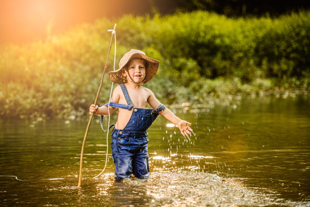 Blonde hair blue toddler smiles with joy as his overall falls off his shoulder. His fishing hat on his head and pole in hand as he splashes creek water during the boys fishing photo session at golden hour.