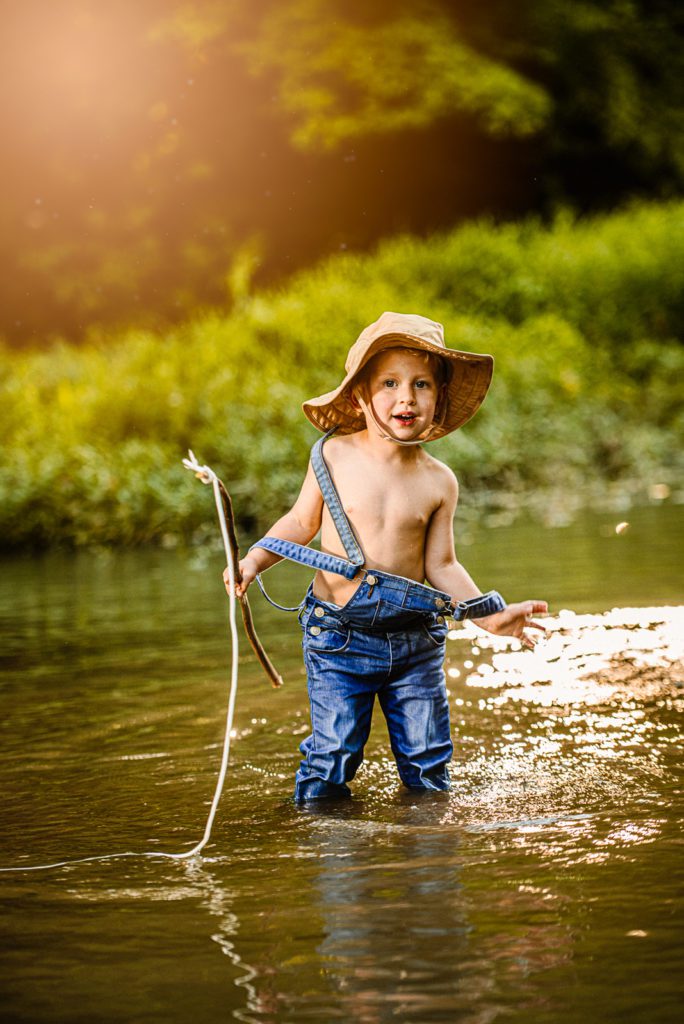 Blonde hair blue toddler smiles with joy as his overall falls off his shoulder. His fishing hat on his head and pole in hand as he splashes creek water during the boys fishing photo session at golden hour.