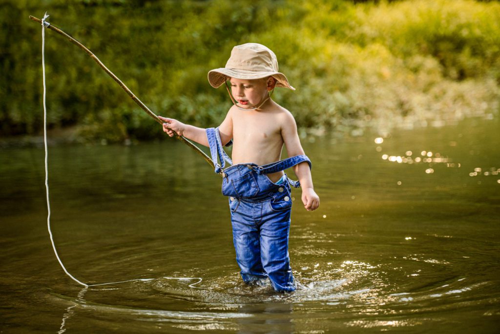Missouri Toddler waits for a fish to take his bait as he stands in the creek dripping with water from splashing in his jeans and hat