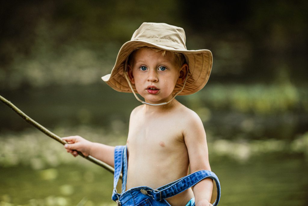 Toddler looks at the camera as the fish got away, overalls falling off his shoulders and hat flopping over his ears.