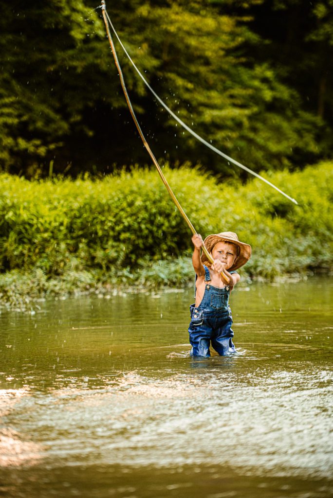 Young boy casts his flying fishing line into a Missouri Creek while wading knee deep in the water surrounded by bright greenery and wearing blue jean overalls and a fishing hat.