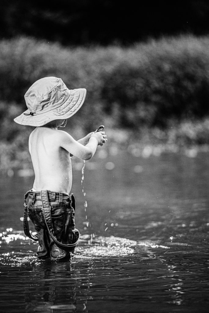Black and white image of toddler grabbing a rock out of the St charles county creek. His overall straps hang at his and water splashes down his elbow