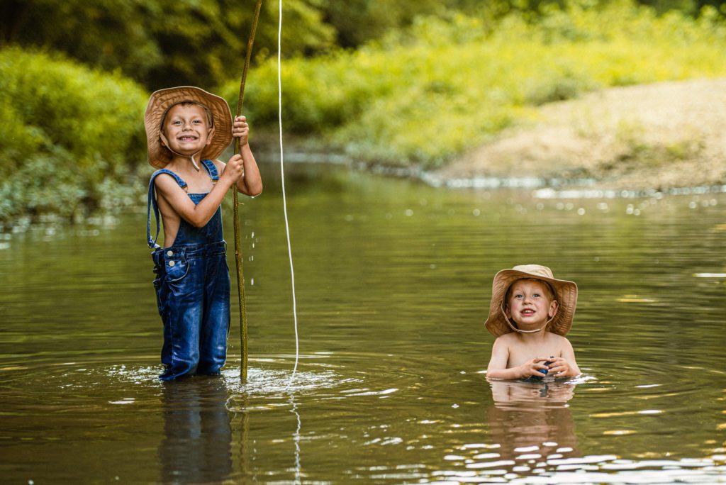Two brothers out fishing in the creek. Older brother stands smiling in his overalls and hat holding his fishing pole high proudly while little brother sits in the creek and plays with a rock and smiles for the camera creating a priceless photography portrait
