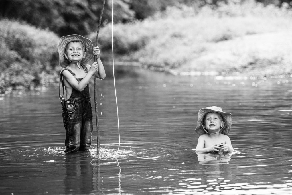 Two brothers out fishing in the creek. Older brother stands smiling in his overalls and hat holding his fishing pole high proudly while little brother sits in the creek and plays with a rock and smiles for the camera creating a priceless photography portrait in black and white.